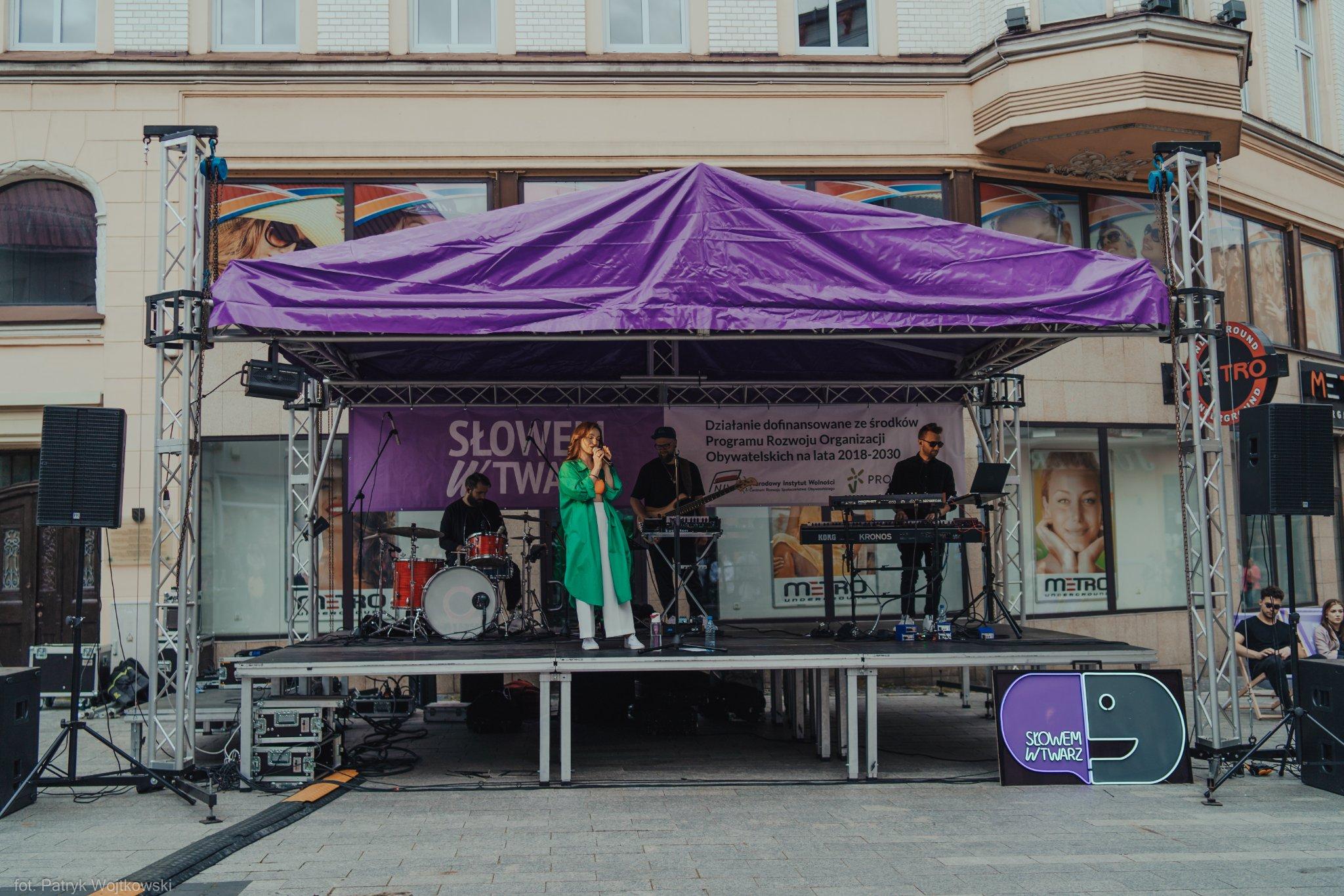 Project concert stage with a band and a "Słowem w twarz" banner during an outdoor event.
